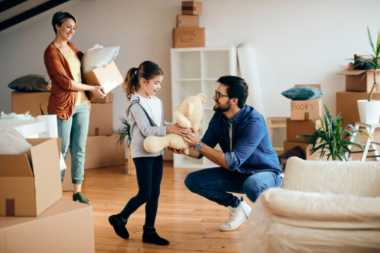 Happy father and daughter having fun with teddy bear while moving into a new apartment. Mother is in the background.